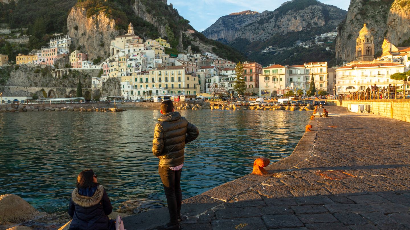 Amalfi's waterfront and hillside buildings across the harbor in late-December light, with two bundled visitors on the stone pier