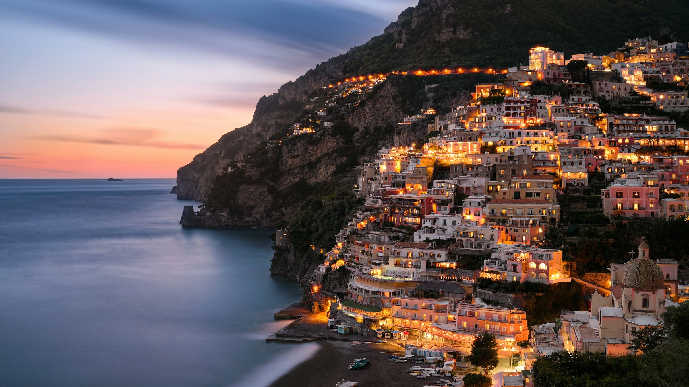 Positano's illuminated hillside buildings above a blue sea beneath a pink and blue evening sky