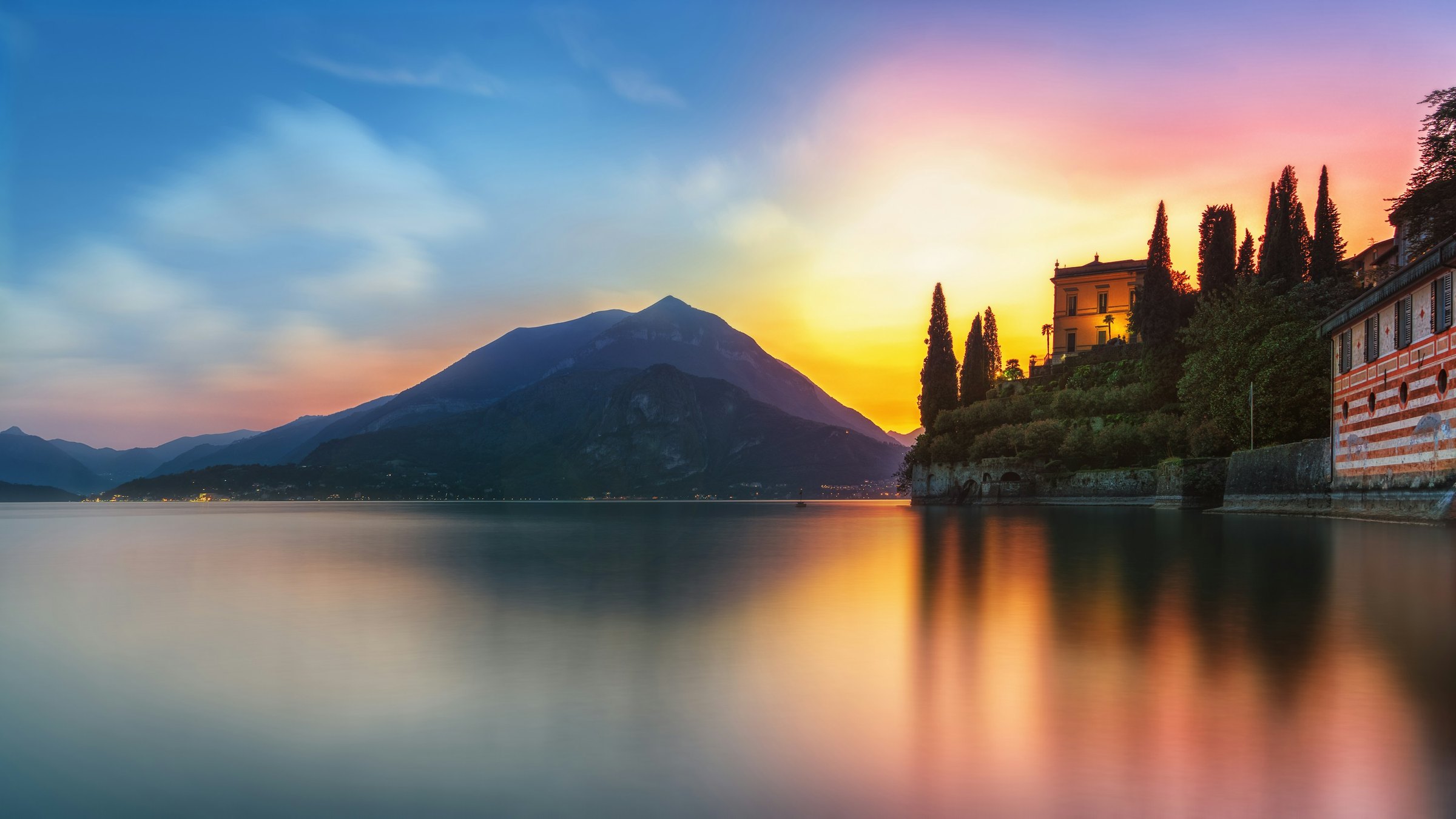 Lake Como and dark mountain layers beneath a blue, gold and pink sunset, with waterfront buildings at the edge