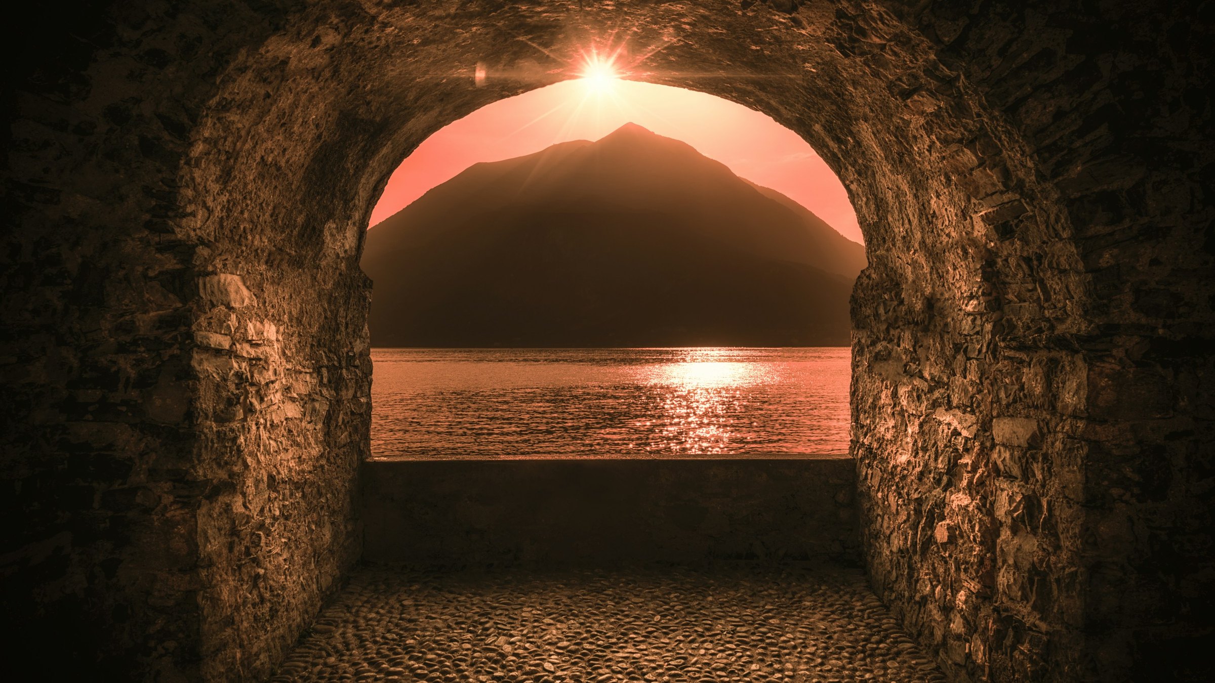 Golden sunset over Lake Como and a mountain silhouette framed by a textured stone archway