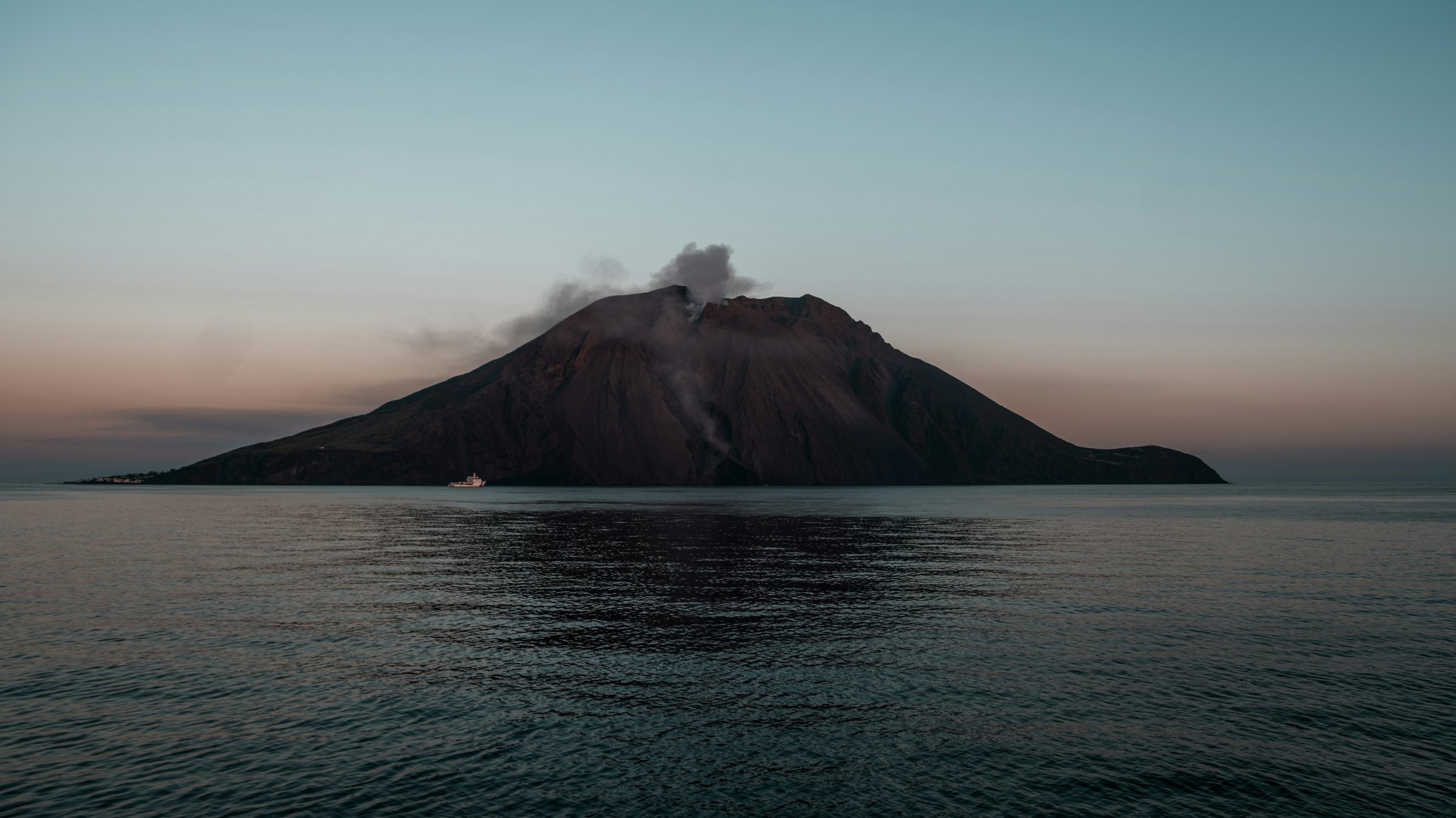 Stromboli's dark volcanic silhouette and a plume above the crater against a soft blue and pink dusk sky, seen across the sea