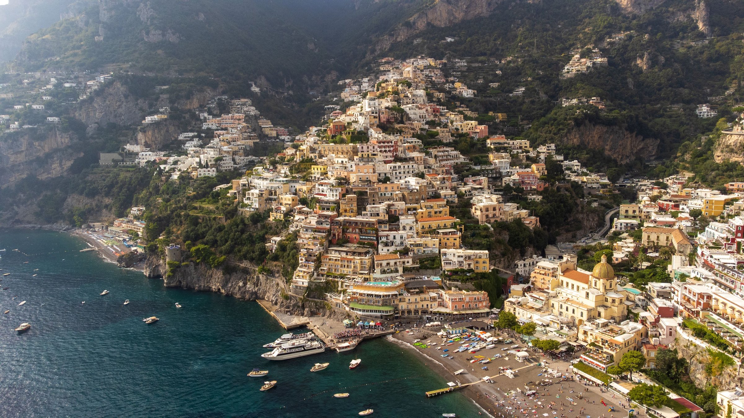 Sunlit Positano climbing a steep wooded coast above deep blue water, with the church dome, beach and boats below
