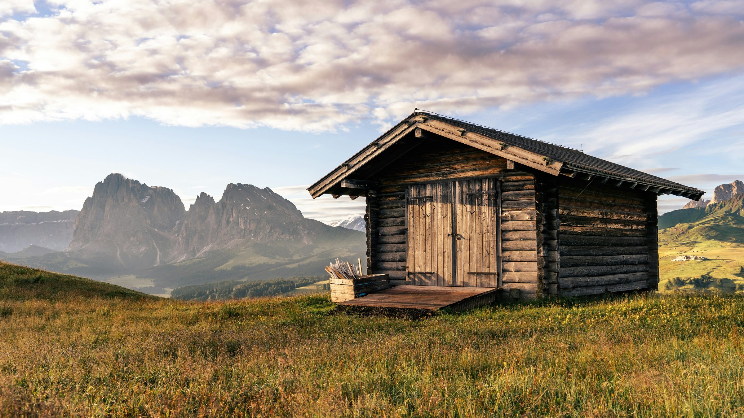 Sunrise over green Alpe di Siusi meadows, a wooden pasture hut and distant Dolomite peaks