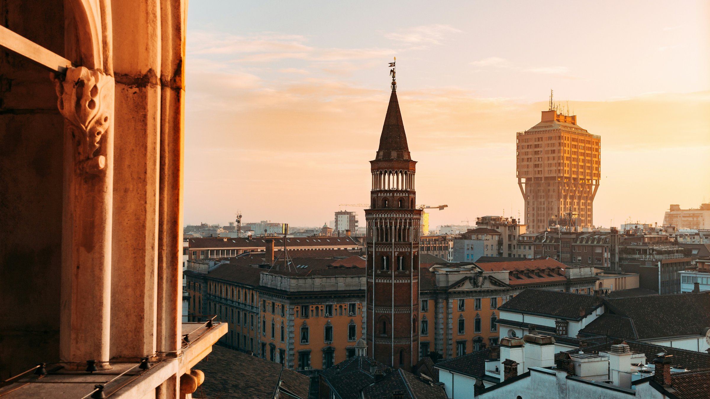 Milan rooftops and towers in golden evening light, framed by a cathedral column