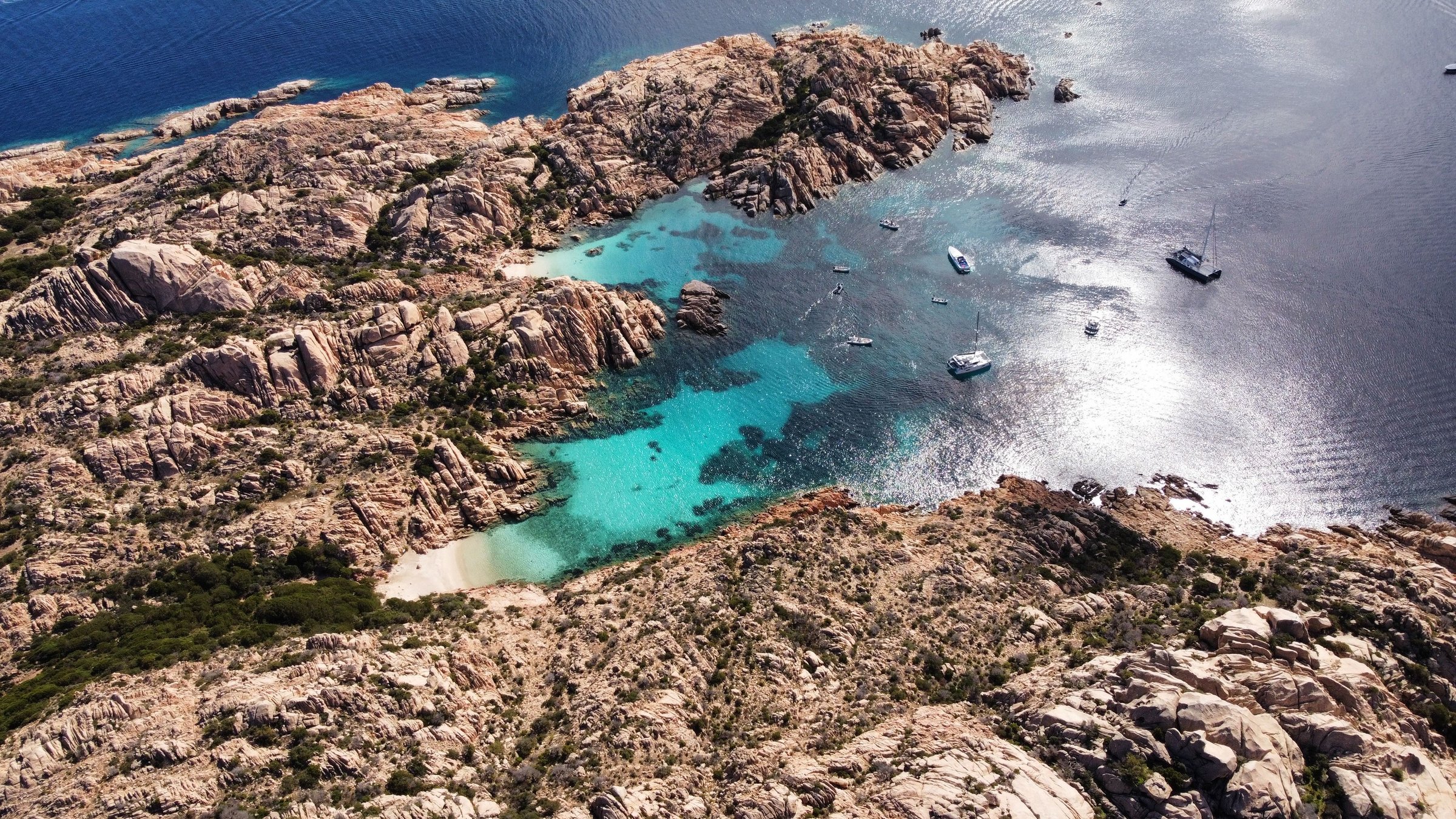 Sculpted granite around turquoise coves in the La Maddalena archipelago, with boats and sunlight on deep blue water
