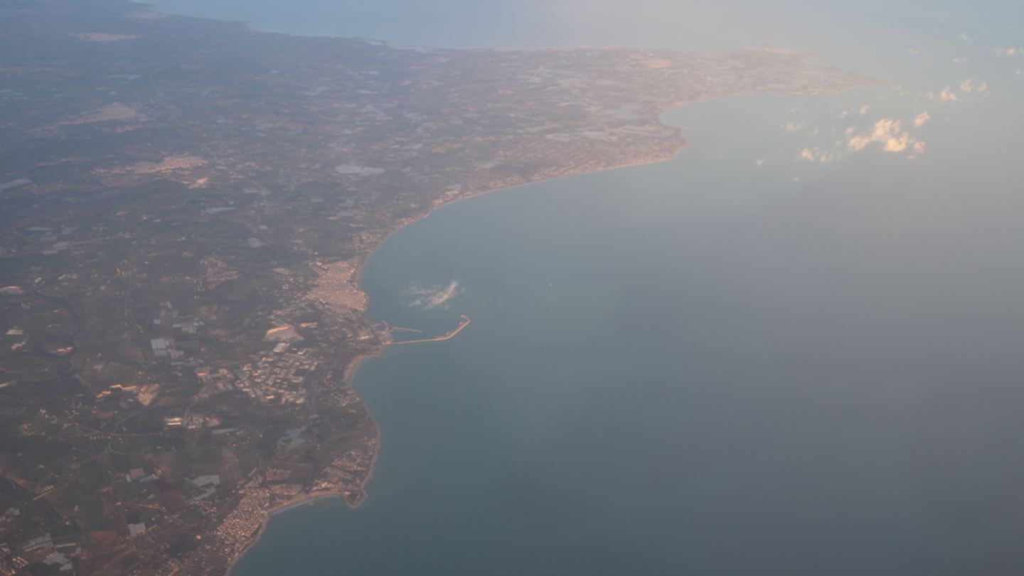 An aerial view across the dry, patterned landscape of Sicily beneath a broad blue sky
