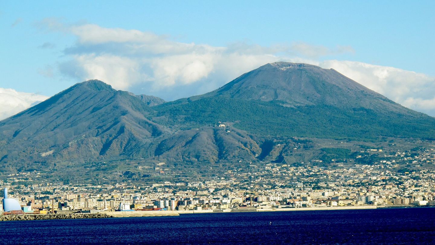 Vesuvius rising above coastal buildings and blue water under a partly cloudy sky